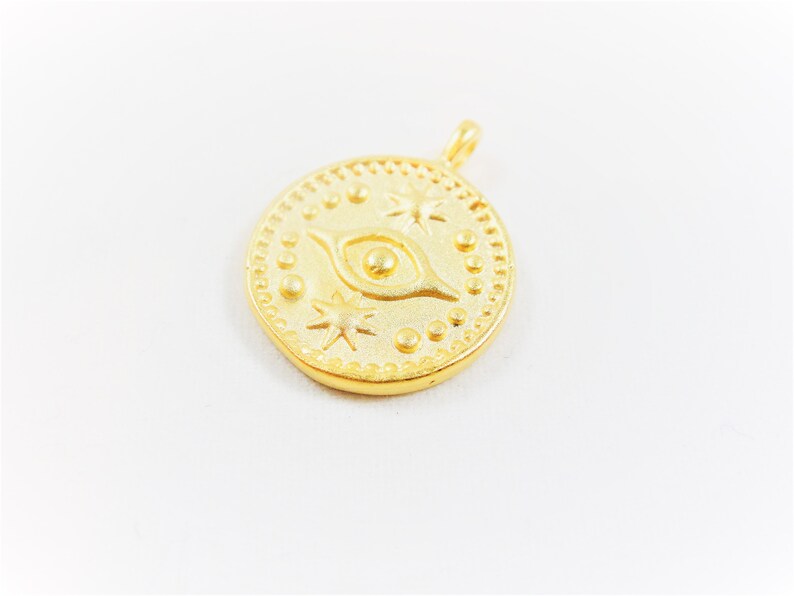 May include: Gold-toned pendant charm featuring a stylized eye with a small circle in the center, surrounded by stars and dots. The charm is round and has a textured surface.