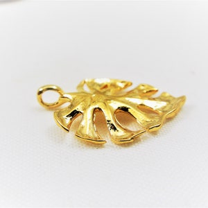 May include: A gold-colored metal pendant shaped like a monstera leaf. The pendant has a loop at the top for attaching to a chain.