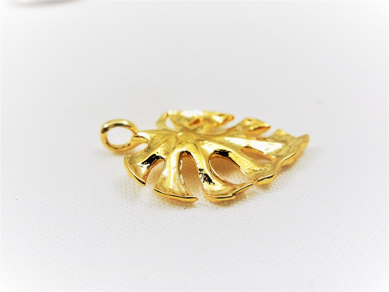 May include: A gold-colored metal pendant shaped like a monstera leaf. The pendant has a loop at the top for attaching to a chain.