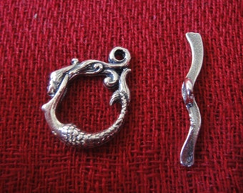 May include: A silver-toned mermaid toggle clasp with a curved bar. The clasp features a detailed mermaid design with a loop for attachment. The bar is wavy with a small loop. The clasp is set against a red fabric background.