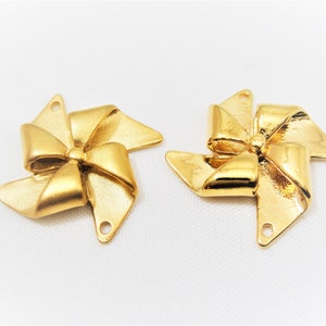 May include: Two gold-colored metal charms shaped like pinwheels with a bow in the center. The charms have a hole at the top for attaching to a necklace or bracelet.