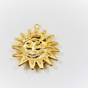 May include: Gold-toned pendant charm in the shape of a sun with a face. The sun has a smiling face with closed eyes and a small nose.