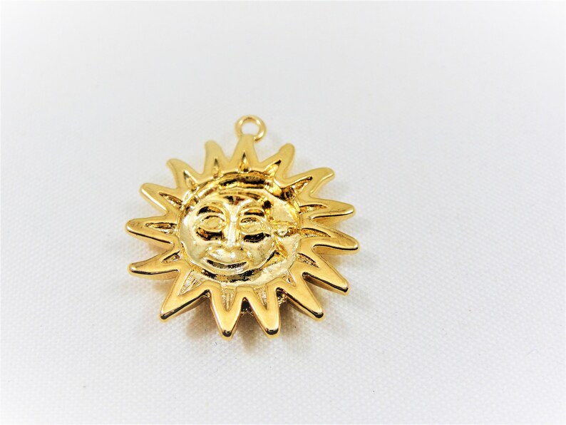 May include: Gold-toned pendant charm in the shape of a sun with a face. The sun has a smiling face with closed eyes and a small nose.