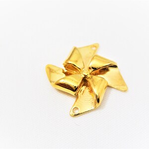 May include: Gold-colored metal charm shaped like a bow with a pinwheel design. The charm has two holes for attaching to a necklace or bracelet.