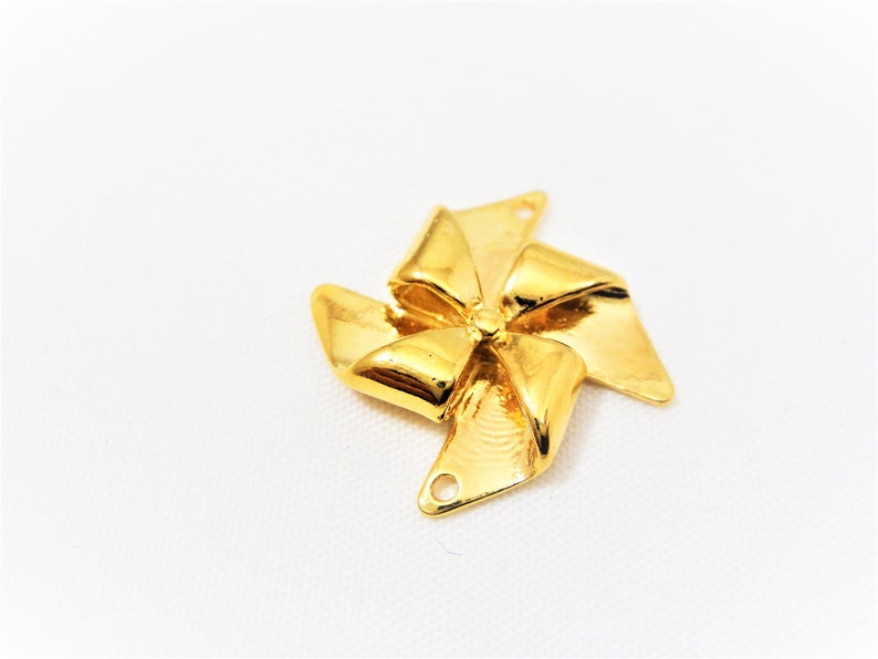May include: Gold-colored metal charm shaped like a bow with a pinwheel design. The charm has two holes for attaching to a necklace or bracelet.