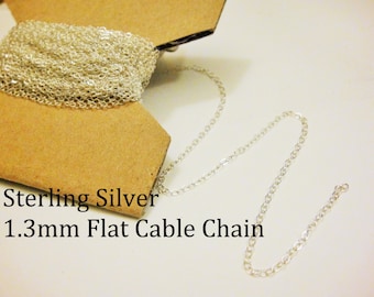 May include: A close-up of a silver 1.3mm flat cable chain. The chain is coiled on a cardboard spool and a section of the chain is laid out on a white surface. Sterling Silver 1.3mm Flat Cable Chain.