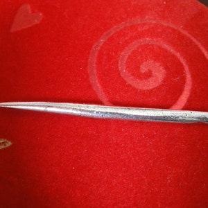 May include: A silver-colored needle with a pointed tip and a small loop at the end. The needle is lying on a red velvet surface.