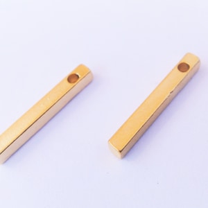 May include: Two gold-colored metal bar pendants with a hole at the top for attaching to a chain or cord.