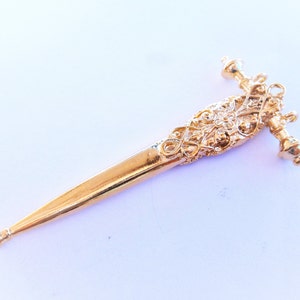 May include: A gold-colored, ornate, decorative dagger with a pointed tip and intricate details. The dagger has a floral design and a rounded handle.