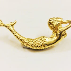 May include: A gold-toned mermaid charm with a detailed design. The mermaid is lying on her side with her tail curled up. The charm has a loop at the top for attaching to a necklace or bracelet.