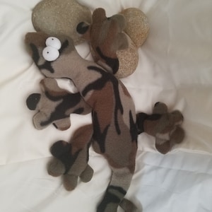 May include: A camouflage-patterned plush gecko toy with large white eyes. The gecko is lying on a white surface.
