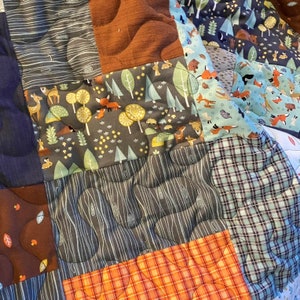 May include: A patchwork quilt featuring various fabric patterns. The quilt includes squares with forest animal prints, tree designs, and plaid patterns. Colors include brown, orange, gray, and blue. The quilt is stitched with a wavy pattern.