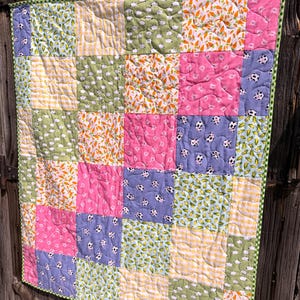 May include: A patchwork quilt featuring a grid of square fabric panels with various patterns including orange carrots, white sheep on green, pink solids, yellow gingham, and purple cow prints, all finished with a green and white checkered border.