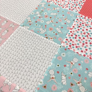 May include: A patchwork quilt featuring alternating square fabric panels. The design includes light blue squares with white rabbits and pink flowers, white squares with a grey swirling line pattern, and white squares with small red and blue flowers.