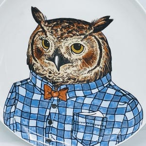 May include: A white ceramic plate featuring a detailed illustration of an owl wearing a blue and white checkered button-down shirt with a small orange bow tie and a pocket square. The owl has brown and black feathers with large yellow eyes.