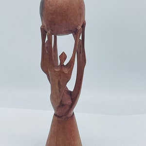 May include: A wooden sculpture of a person holding a sphere above their head. The sculpture is made of dark brown wood and has a simple, abstract design.