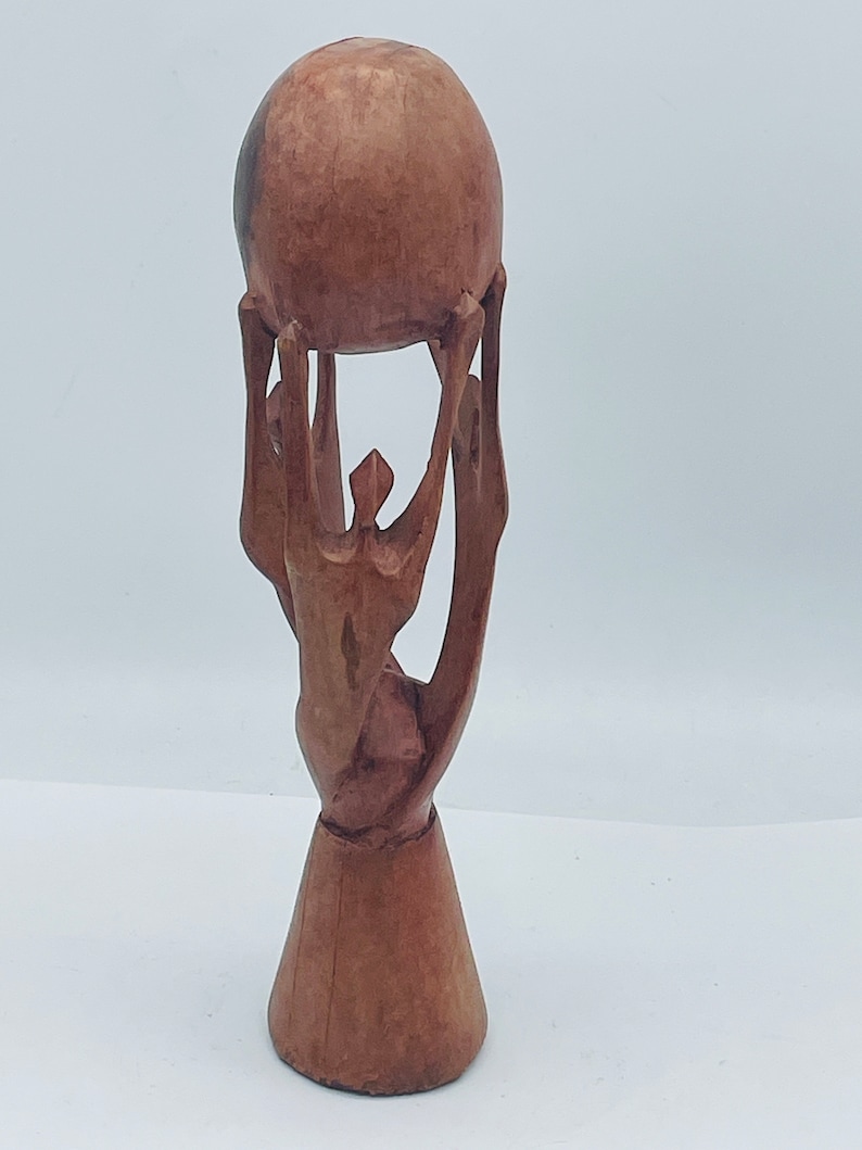 May include: A wooden sculpture of a person holding a sphere above their head. The sculpture is made of dark brown wood and has a simple, abstract design.