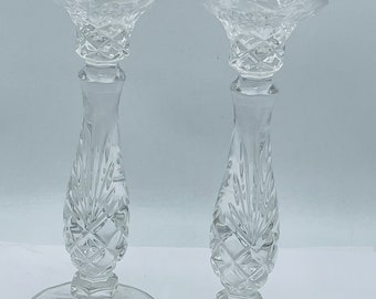 May include: A pair of clear glass candle holders with a cut crystal design. The holders have a wide base and a tall, slender stem. The top of each holder is a wide, open cup for holding a candle.