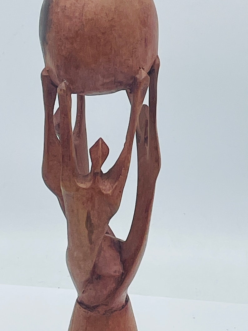 May include: A wooden sculpture of a person holding a sphere above their head. The sculpture is made of dark brown wood and has a smooth, polished finish.