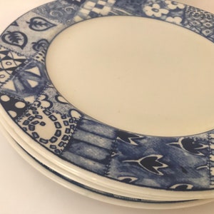 May include: Two white ceramic plates with a blue and white patterned border. The pattern features a patchwork design with various geometric shapes and floral motifs.