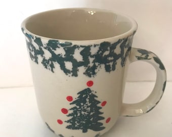 May include: Cream-colored ceramic mug with a dark green Christmas tree design and red ornaments. The mug has a dark green border around the rim and a rounded handle. A festive holiday mug.