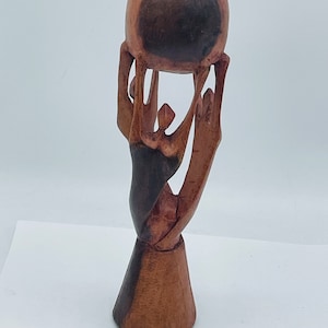 May include: A wooden sculpture of three figures holding up a sphere. The sculpture is made of dark brown wood and has a natural finish.