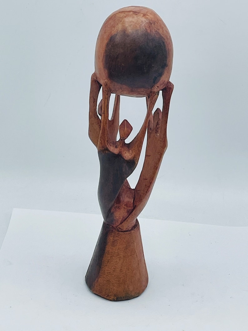 May include: A wooden sculpture of three figures holding up a sphere. The sculpture is made of dark brown wood and has a natural finish.