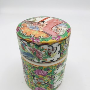May include: A round porcelain container with a lid, featuring a colorful floral and figural design. The lid depicts a scene with figures in traditional clothing, while the body of the container is adorned with a vibrant floral pattern.