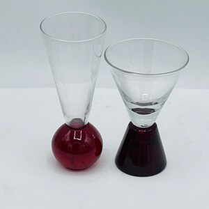 May include: Two clear glass vessels with distinct bases. The left vessel features a tall, slender, conical bowl atop a spherical red glass base. The right vessel has a wider, flared bowl sitting on a solid, truncated conical red glass base.