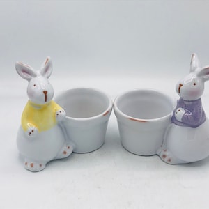 May include: Two white ceramic bunny egg cups. One bunny is wearing a yellow sweater and the other is wearing a purple sweater.