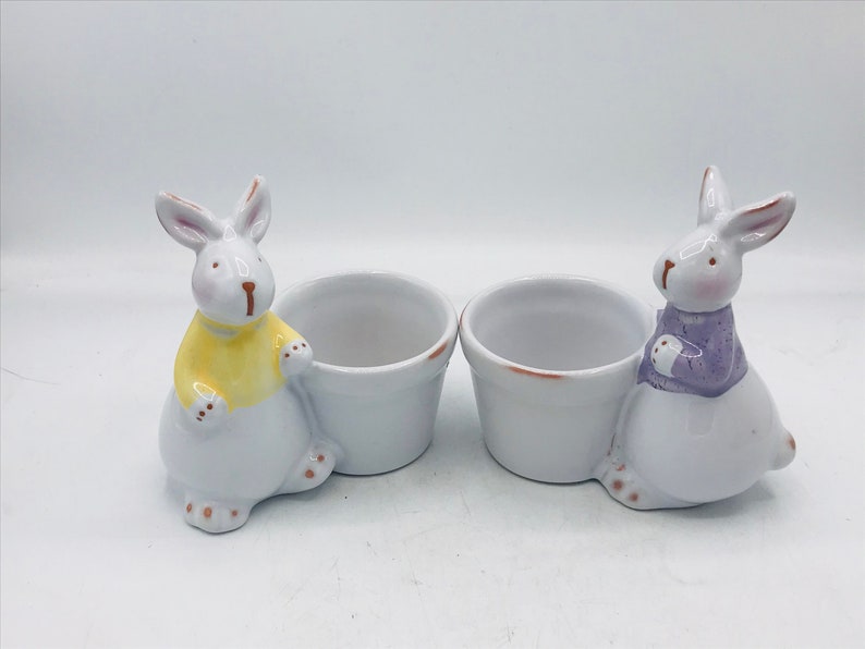 May include: Two white ceramic bunny egg cups. One bunny is wearing a yellow sweater and the other is wearing a purple sweater.
