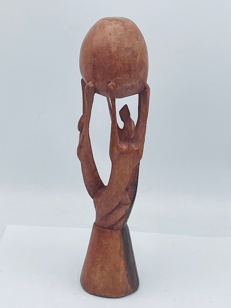 May include: A wooden sculpture of three figures holding up a sphere. The figures are stylized and abstract, with long, thin limbs. The sphere is smooth and round, and it is slightly larger than the figures' heads. The sculpture is made of a dark brown wood and has a natural finish.
