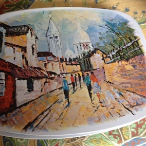 May include: A white ceramic dish with a painted scene of a Parisian street. The scene includes buildings, a church, and people walking down the street. The painting is in a colorful, impressionistic style.