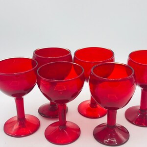 May include: Set of six red glass wine glasses with a rounded bowl and a short stem. The glasses are arranged in a circle on a white surface.