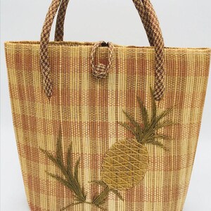 May include: Woven tote bag with a pineapple design. The bag features a rectangular shape with a woven pattern in shades of beige and brown. It has two braided handles and a decorative pineapple applique.