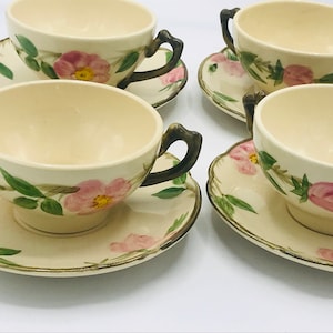 May include: Set of four vintage teacups and saucers with a floral pattern. The cups have a cream-colored base with a pink rose design and green leaves. The saucers have a matching design. The cups have gold-colored handles.