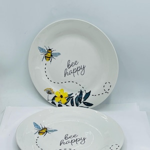May include: Two white ceramic plates with a bee and floral design. Each plate features a yellow and black bee, yellow flowers, and the phrase "bee happy" in black script. The plates have a simple, elegant design.