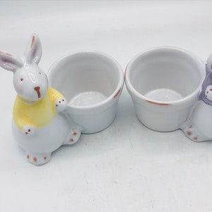 May include: Two white ceramic bunny egg cups. One bunny is wearing a yellow shirt and the other is wearing a purple shirt.