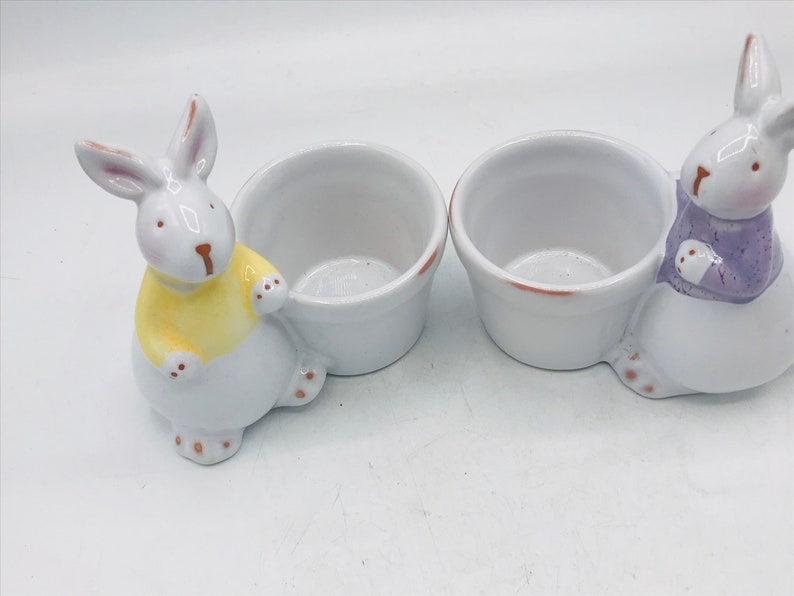 May include: Two white ceramic bunny egg cups. One bunny is wearing a yellow shirt and the other is wearing a purple shirt.
