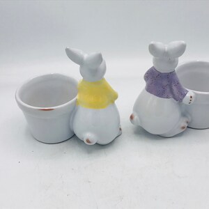 May include: Two white ceramic bunny planters with small pots. One bunny is wearing a yellow sweater and the other is wearing a purple sweater.
