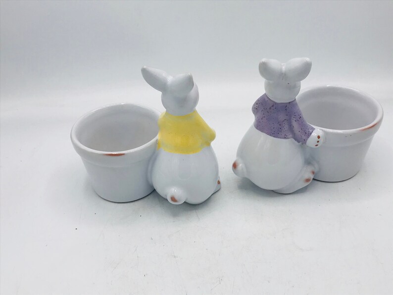 May include: Two white ceramic bunny planters with small pots. One bunny is wearing a yellow sweater and the other is wearing a purple sweater.