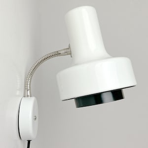 May include: A white wall-mounted lamp with a flexible, silver-colored gooseneck arm. The lamp head is white with a black underside. The lamp is mounted on a white wall.