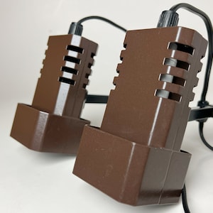 May include: Two brown, rectangular spotlights with a slotted design, connected by black wires and mounted on a black bracket. The lights are angled towards the viewer, showcasing their geometric shape and the adjustable knob.