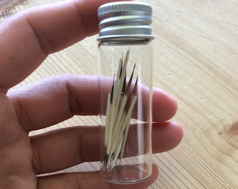 Puede incluir: Un pequeño frasco de vidrio con tapa plateada que contiene una colección de plumas blancas delgadas.