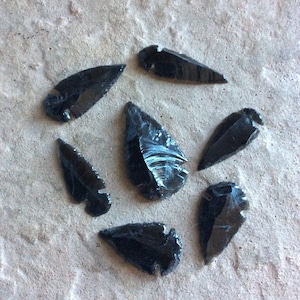 May include: Seven black obsidian arrowheads, also known as obsidian points, on a light gray textured surface.