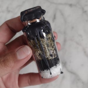 May include: A small clear glass vial sealed with a cork and topped with black wax that drips down the sides. The interior is filled with distinct layers of white salt, black sand, dried herbs, and light-colored botanical pieces in a vertical stack.