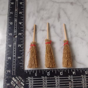 May include: Three miniature brooms with wooden handles and straw bristles, each secured with a red twine. The brooms are arranged on a surface next to a ruler.