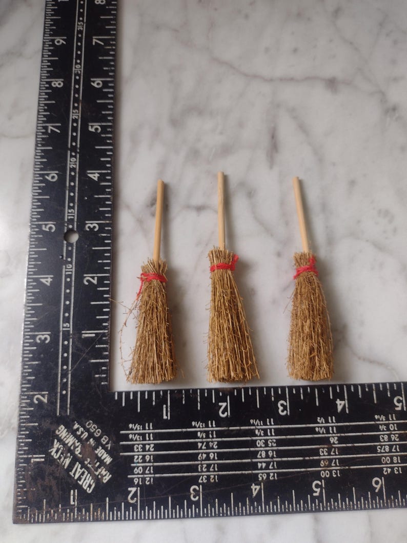 May include: Three miniature brooms with wooden handles and straw bristles, each secured with a red twine. The brooms are arranged on a surface next to a ruler.