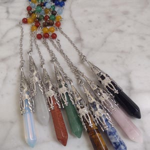 May include: A collection of crystal pendulums in various colors, including blue, pink, green, and black. Each pendulum has a silver-colored metal cap and chain, with a beaded chain at the top. The crystals are displayed on a white marble surface.