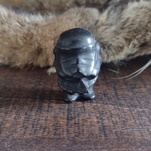 May include: A small, black, carved figurine of a person with a helmet-like head and a detailed face. The figurine is standing on a wooden surface.